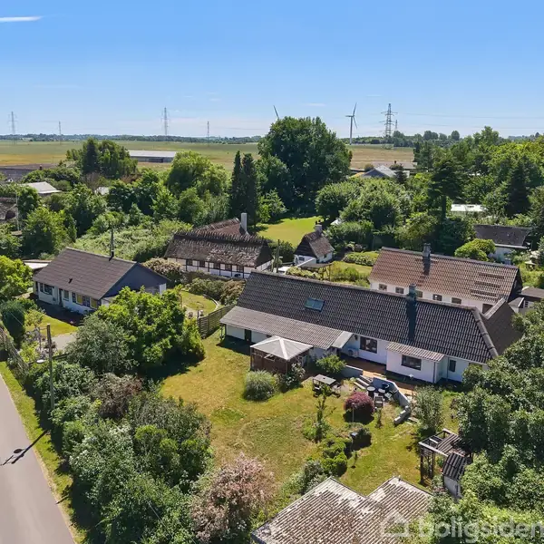 Huse ligger langs en vej omgivet af frodig grønne haver og træer, med åbne marker og vindmøller i baggrunden under en klar blå himmel.