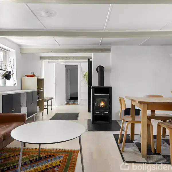 Stue med brændeovn i centrum, omgivet af en brun lædersofa, rundt hvidt sofabord og træspisebord med stole. Rummet har hvide vægge og gulvtæpper, med naturligt lys fra vinduer.