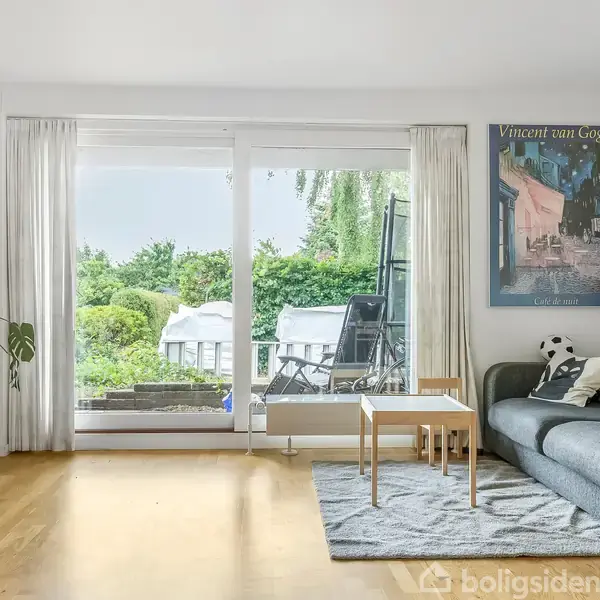 Stue med sofa og sofabord foran et stort vindue. På væggen hænger en plakat med teksten "Vincent van Gogh". Udenfor ses grøn have og havemøbler.