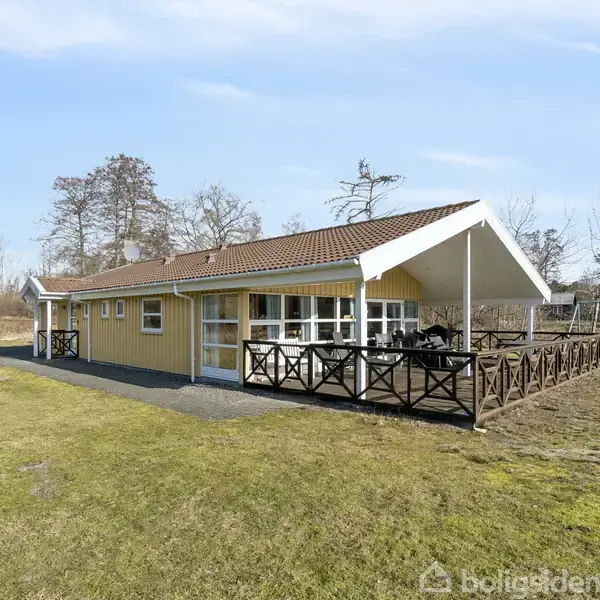 Gult sommerhus med terracotta-tag, står stille omgivet af græs og spredte træer. Der er en stor træterrasse med rækværk foran huset.