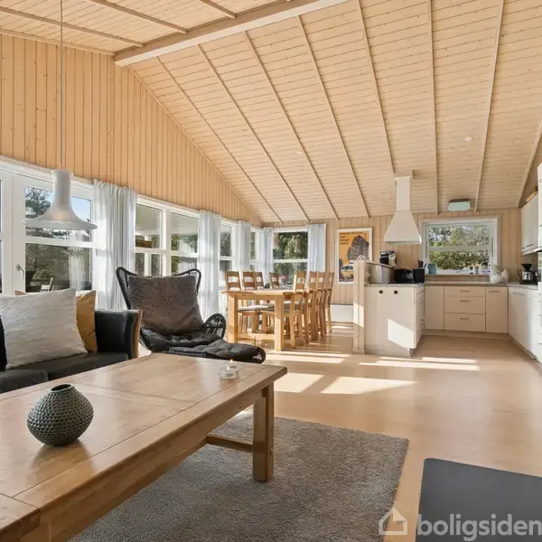 Stue med træbeklædte vægge og loft. Sofa og sofabord foran pejsen. Spisebord og køkken i baggrunden, belyst af store vinduer.