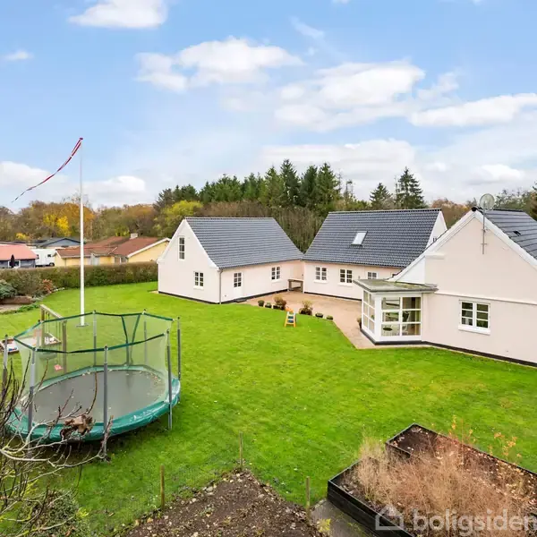 Et stort, moderne hus med gråt tag står roligt i en velholdt grøn have med trampolin og flagstang. Skov omkranser ejendommen i en fredelig forstadsidyl.