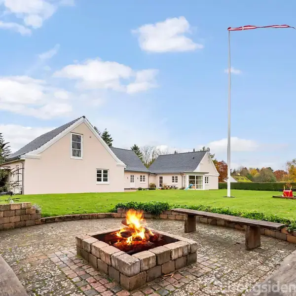 Bål i en firkantet stenrand brænder i en gård med græsplæne. Hvidt hus med mørkt tag ses i baggrunden. Flagstang og trampolin er til stede.