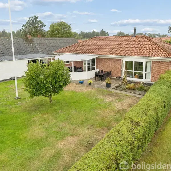 Et rødt hus med tegltag står i en grøn have med omkransende hæk. Foran er en terrasse med havemøbler, og et flagstang står til venstre. I baggrunden ses et hvidt hus.