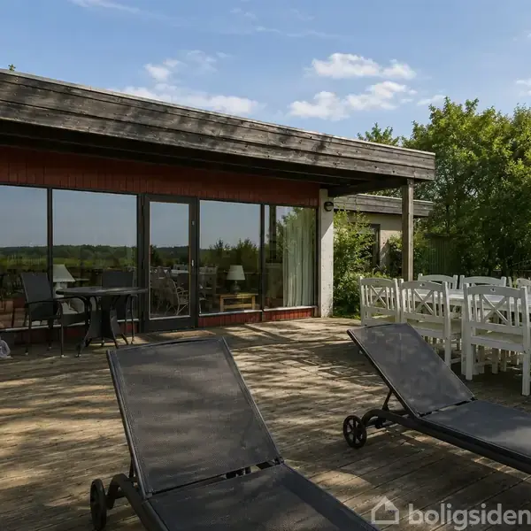 Terrasse med to sorte liggestole foran et moderne hus med store glasdøre. En stor spisebordssæt står til højre. Omkringliggende grønt landskab og blå himmel skaber en fredelig stemning.