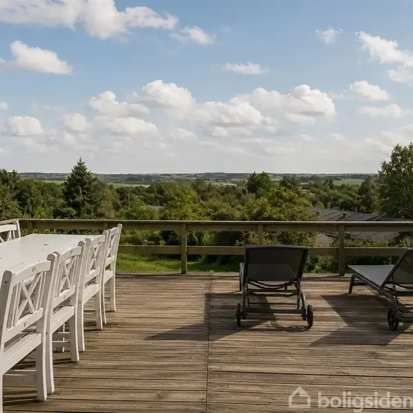 På en terrasse står et langt hvidt spisebord med otte stole ved siden af to sorte liggestole. I baggrunden ses grønne træer og en blå himmel med skyer.