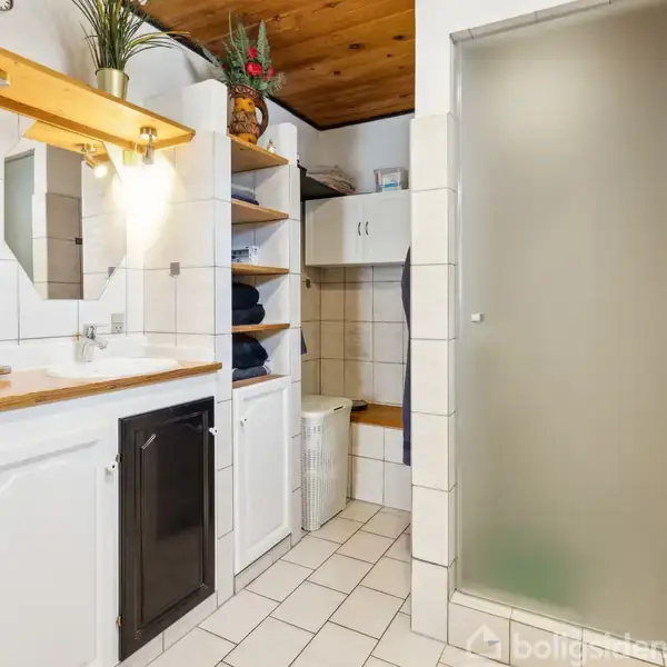 Badeværelse med hvide fliser, hvidt skab med sorte låger, brusekabine med matteret glas og hylder med håndklæder. På træhylde står planter. Flisegulv og vægmonteret håndklædetørrer.