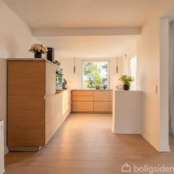 Et lyst køkken med trægulv, hvor en enkel plante og blomsterdekoration pryder skabene. Store vinduer lader naturligt lys strømme ind, og gardiner hænger til højre.