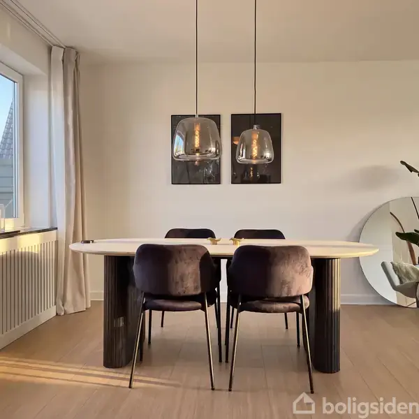 Et moderne spisebord med fire mørke stole står i en lys spisestue. To hængelamper svæver over bordet. Store vinduer og spejl tilføjer lys; planter pynter hjørnet.