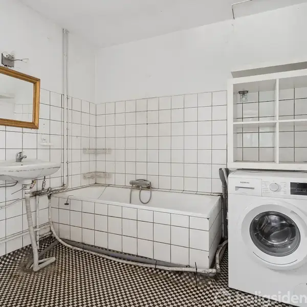 Badekar og vaskemaskine i et badeværelse med hvide fliser på væggene og sort-hvidt ternet gulv. Vask med spejl hænger over en radiator. Skab monteret på væggen.