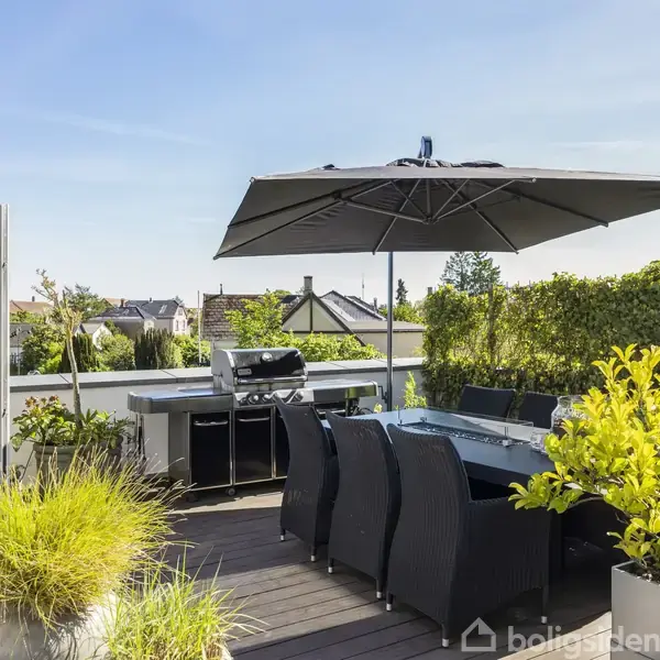 Terrasse med spisebord og stole under en stor parasol. I baggrunden ses en grill og grønne planter, der omkranser området med udsigt til huse og blå himmel.