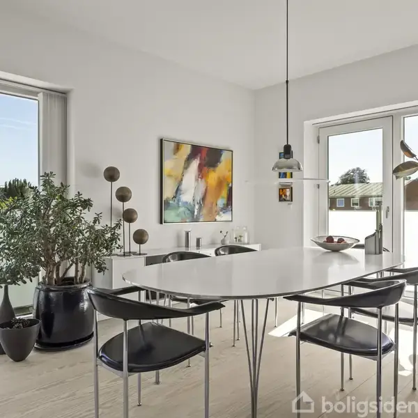 Et moderne spisestuemøblement står i et lyst rum med glasdøre, omgivet af grønne planter og moderne kunst på væggen, hvilket skaber en stilfuld, indbydende atmosfære.