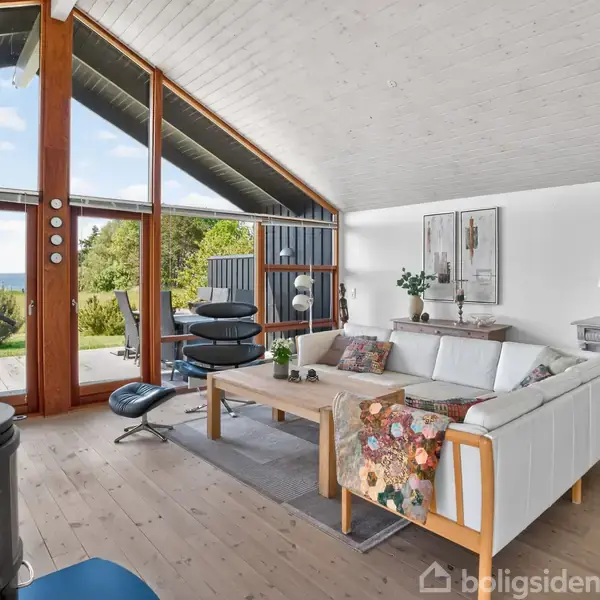 Stue med hvid sofa og lænestol, der vender mod store vinduer med udsigt til en grøn have. Trægulv og -loft skaber en lys og hyggelig atmosfære. Pejs i forgrunden.
