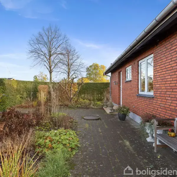 Rød murstensbygning står stille, omgivet af en have med bare træer og efterårsløv. En træbænk med græskar og planter står tæt ved husets murstensvæg under en klar blå himmel.