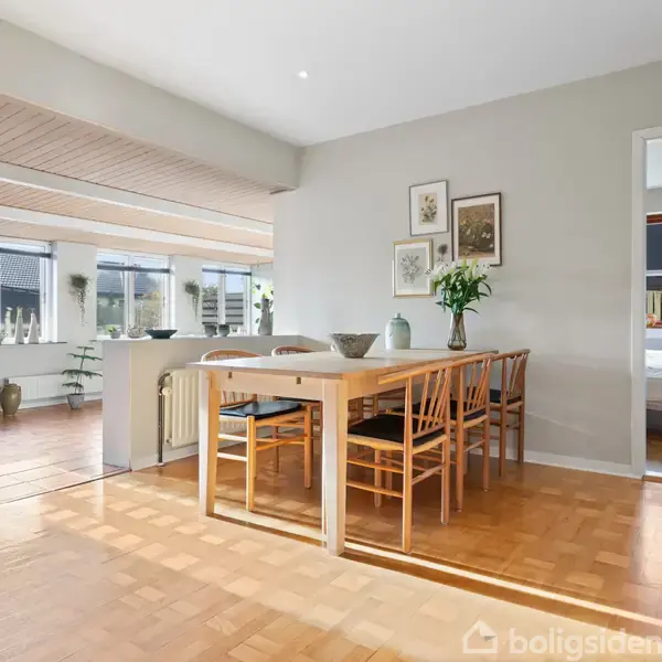 Et spisebord med seks træstole står i en lys spisestue. På væggen hænger billeder, og en stor vase med blomster står på bordet. Rummet har parketgulv og store vinduer.