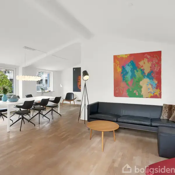 En moderne stue med en sort sofa og farverigt maleri på væggen. Et rundt sofabord står foran sofaen. Til venstre er en spiseplads med stole og stor lampe.