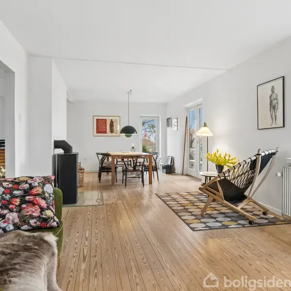 Sofa med blomstrede puder står i en lys stue med trægulv. Spisebord og stole ses i baggrunden. Vægkunst og vindue med udsigt til have.