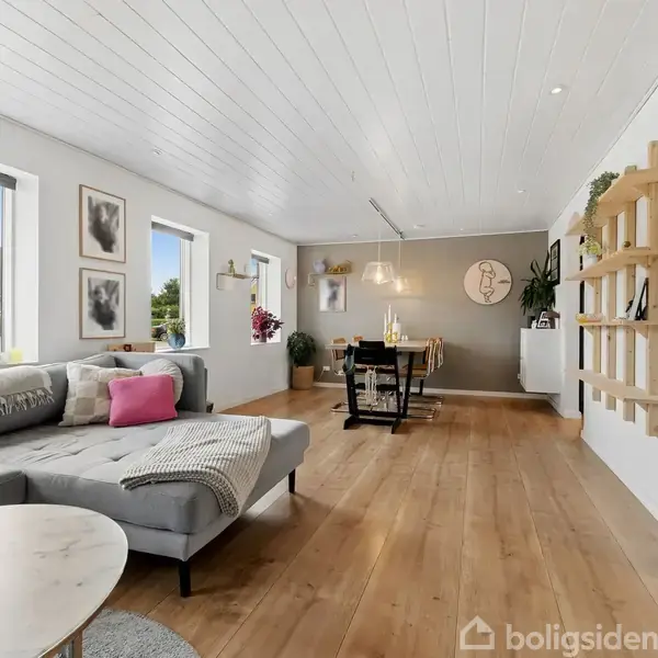 En lys stue med en grå hjørnesofa og et rundt sofabord i forgrunden. Fjernere ses en spiseplads med et bord, stole og vægdekoration i et rummeligt og moderne hjem.