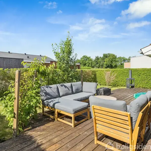 Terrassemøbler er arrangeret på en træterrasse med grå sofahjørne og træstole. Terrasse omgivet af grøn hæk med udsigt til nabobygninger under en blå himmel og sollys.