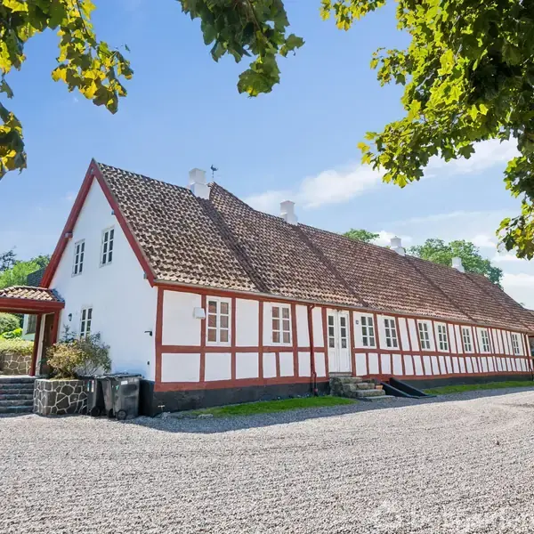 En hvidkalket bindingsværksbygning med rødt tag står stille på en grusbelagt gårdsplads, omgivet af grønne træer og klar blå himmel.
