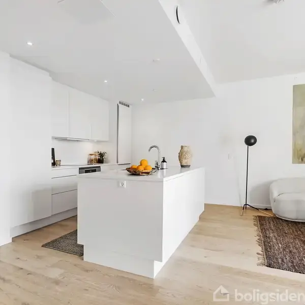 Et moderne køkken med en hvid ø, fyldt med frugter og dekorative genstande, er indrammet af hvide skabe. I baggrunden ses en hyggelig stue med sofa og maleri.