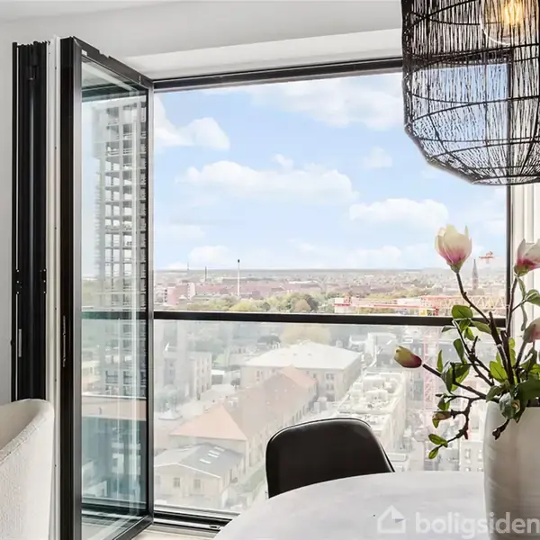 En åben altandør fører ud til en udsigt over byen med bygninger i baggrunden. I forgrunden er der et rundt bord med en vase med blomster.