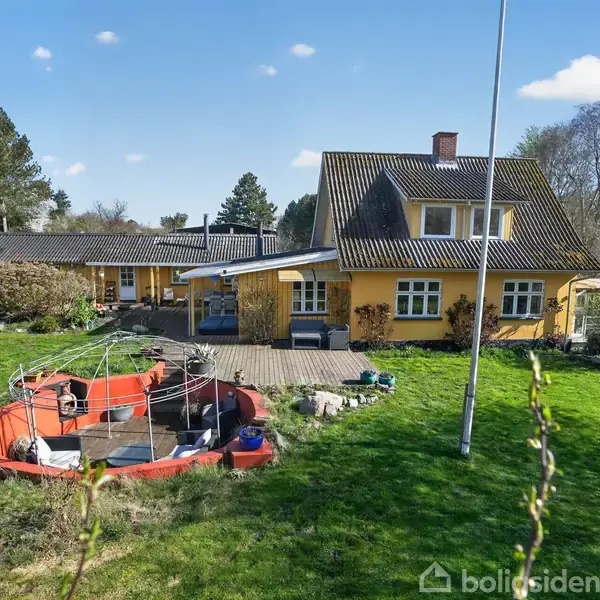 Et gult hus med mørkt tag står i en frodig have. Foran huset er en terrasse, og til venstre ses en båd placeret i græsset.