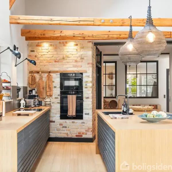 Køkken med træbjælker i loftet og murstensvæg. Køkkenelementerne har træoverflader, og der hænger to lamper over køkkenøen. Planter og køkkenredskaber pynter rummet.