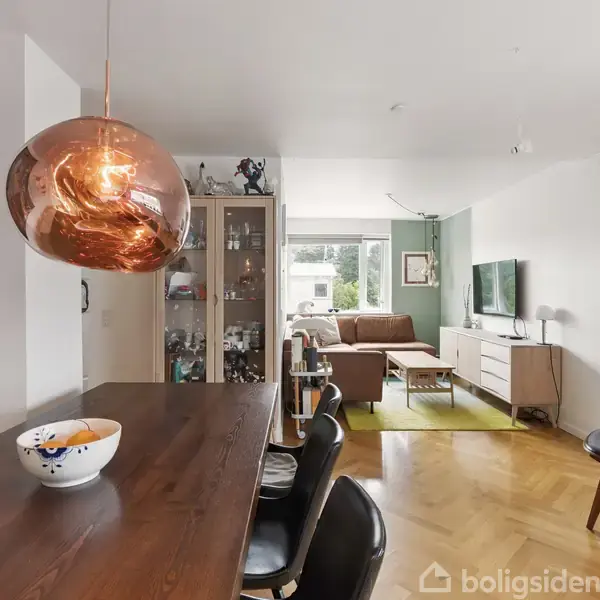 Kobberlampe hænger over et mørkt træbord med en skål. I baggrunden viser en stue med sofa, TV og skab. Lyse vægge og parketgulv skaber en moderne atmosfære.