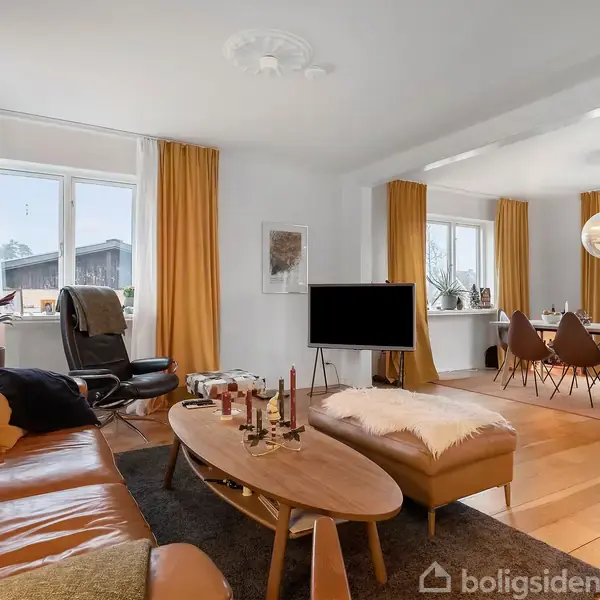 Brun lædersofa står i stue med træbord foran, flankeret af en lænestol. Fjernsyn midt i rummet. Spisebord og stole i baggrunden, omringet af hvide vægge og gule gardiner.
