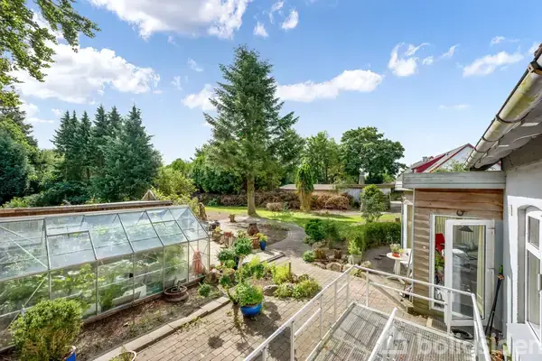 Et drivhus står på en terrasse, omgivet af grønne planter og grusstier. I baggrunden ses en velplejet have med høje træer under en skyet blå himmel.