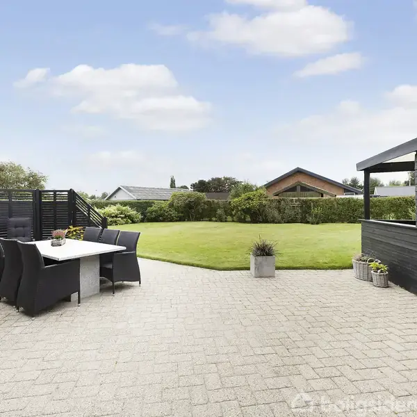 Et rødt murstenshus med sort tag står i en stor, grøn have. Foran ses en terrasse med hegn. Himlen er blå med få skyer.