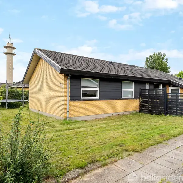 Et gult murstenshus står stille i en veltrimmet have med en flisebelagt gangsti. I baggrunden ses et højt tårn under en blå himmel.