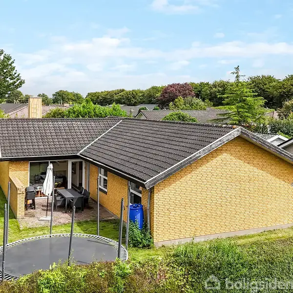 Gul murstensvilla med mørkt tag, omgivet af grøn have med trampolin og terrassemøbler. Beliggende i et stille villakvarter med andre huse og træer i baggrunden.