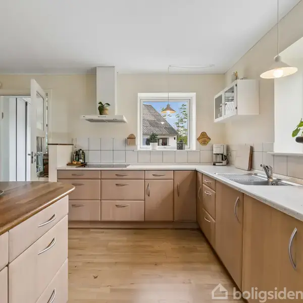 Lyst køkken med træskabe og hvide bordplader. Frisk frugt på køkkenøen. Vindue viser blå himmel og et hus. Indendørs planter står ved håndvasken.