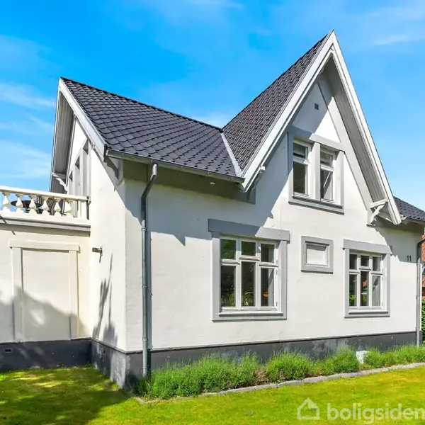 En hvid bungalow står i en frodig have med grøn græsplæne og høje træer omkring. Huset har en mørk tagkonstruktion og flere vinduer.