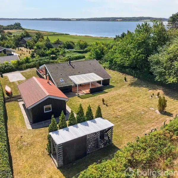 Et sommerhus med terrasse er omgivet af grønne arealer og træer. En mindre bygning står ved siden af, og havudsigt kan ses i baggrunden, hvor også boliger er synlige.