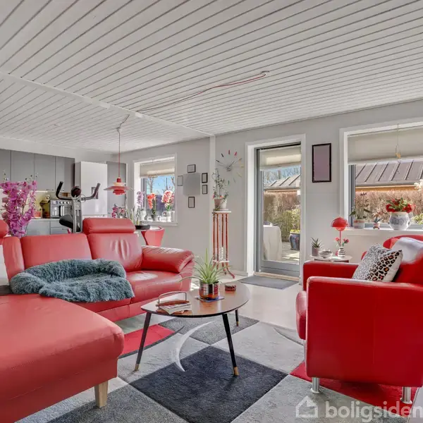 Røde sofaer og en stol er placeret i en moderne stue med lysindfald fra vinduerne. Et sofabord står på et farverigt gulvtæppe, omgivet af dekorative planter og nips.