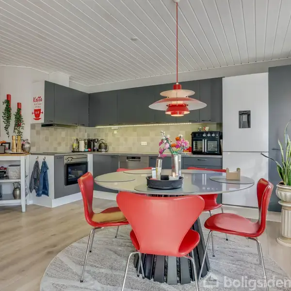 Rundt spisebord med røde stole står under en rød lampe. Køkken med grå skabe og indbygget ovn i baggrunden. Vindue med blomster til højre skaber lysindfald.
