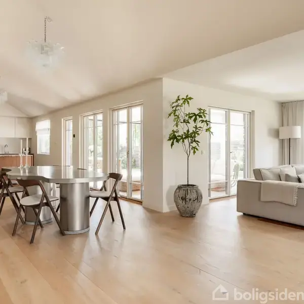 Spisestue med stort, rektangulært bord og seks stole, beliggende ved siden af en stue med grå sofa og stor plante. Rummets lyse trægulv og vinduer skaber en lys atmosfære.