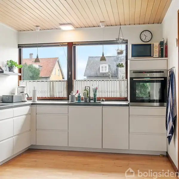 Køkken med hvide skabe og indbygget ovn, har udsyn gennem store vinduer til huse udenfor. Loftet er træ og væggen har en vægur. Der hænger et viskestykke ved siden af ovnen.