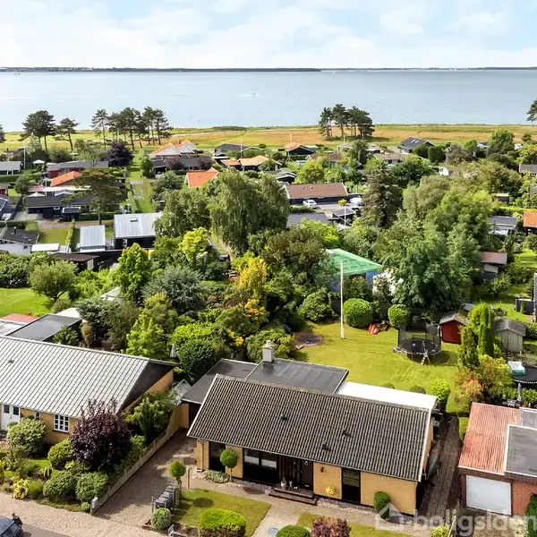 Huse står stille i en grøn, kystnær landsby ved vandet, omgivet af haver og træer. I baggrunden ses kystlinjen og havet.