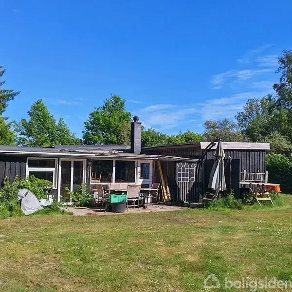 Et sortmalet træhus står stille i en solrig have omgivet af høje træer. Foran huset er der havemøbler og en terrasse, mens en græsplæne breder sig omkring.