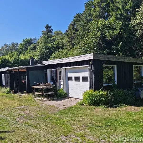 En sort træbygning med garage står stille i en grøn have omgivet af tæt skov. En trailer er placeret ved siden af bygningen, og en træbænk står foran.