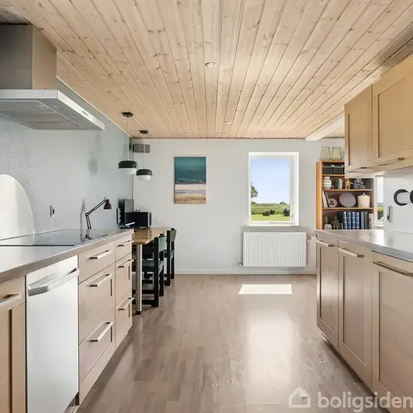 Køkken med træloft og trægulv, indrettet med moderne apparater og skabe på begge sider. Et vindue i baggrunden giver udsigt til grønt landskab. Vægkunst og bøger findes på hylder.