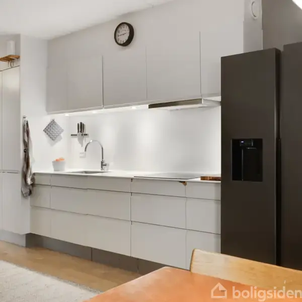 Køkken med moderne grå skabe og en indbygget vask, set i et lyst rum. En sort køleskab står til højre. Vægmonteret ur over skabene. Teppe på gulvet.