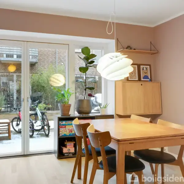 Spisebord med træstole er placeret i en spisestue. Vægge i beige. Store glasdøre fører til en terrasse med cykel. Loftslampe hænger. Grønne planter og kunst dekorerer rummet.