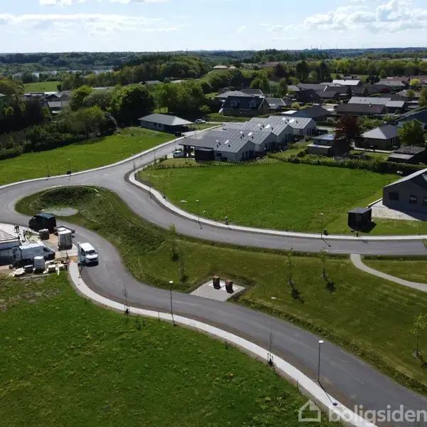 En snoet vej løber gennem et grønt boligområde med parcelhuse og byggerier. Landsbylignende omgivelser med åbne marker og tætliggende huse i baggrunden.