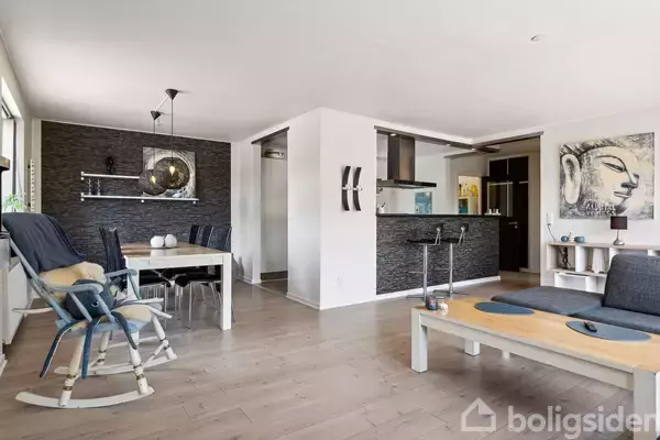 Stue med træmøbler og gyngestol, hvor et spisebord er sat i nærheden af køkkenbaren. Vægge dekoreret med kunst, danner en moderne og stilfuld atmosfære.