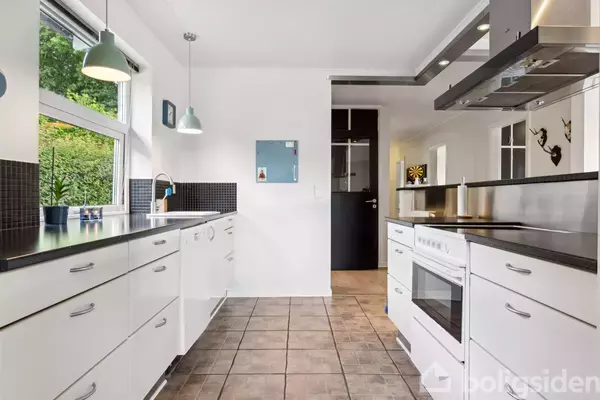 Hvidt køkken med sorte bordplader, ovn og skabe, åbner sig mod en lys gang. Grøn udsigt ses gennem vindue; lamper hænger over arbejdsflade. Fliser dækker gulvet.